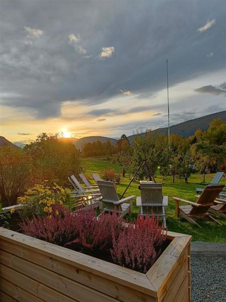 Orheimstunet i Nordfjord - Hagelandskap med trestolar og blomsterkasse i forgrunnen, solnedgang over grøne marker og skyer..