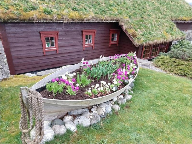 Orheimstunet i Nordfjord - Ein båt er fylt med jord og blomstrar som blomsterbed framfor eit raudt trehus med torvtak og plen..