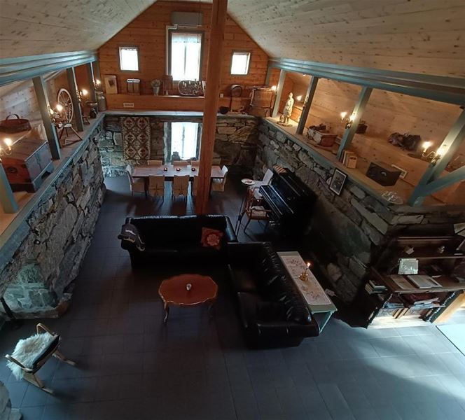 Orheimstunet i Nordfjord - Oversyn frå loftet av ei rustikk stove med steinveggar, sofa, piano, spisebord og tregolv under skråtak..