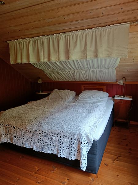 Orheimstunet i Nordfjord - Dobbeltseng med hekleteppe under skrått tretak, med sengekappe over og nattbordlamper på kvar side..