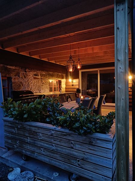 Orheimstunet i Nordfjord - Overbygd terrasse med treverk og steinvegg, grill, bord og stolar, varm lyktelys og plantekasse i front..