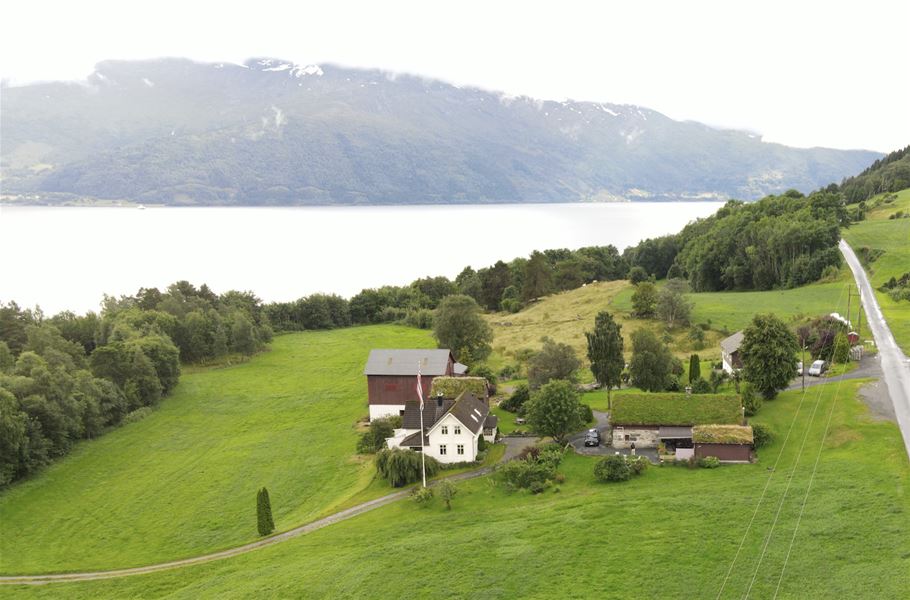 Orheimstunet i Nordfjord - Luftfoto av ein gard med kvitt hus og raud løe, grøne jorde og tre, med fjord og fjell i bakgrunnen..