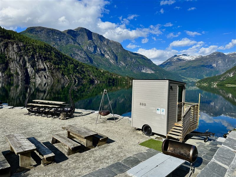 Utesitteplass med bord og benkar ved stille fjord, lita hytte på hjul og fjell under blå himmel –Panorama Sauna ved Strynsvatn Camping..