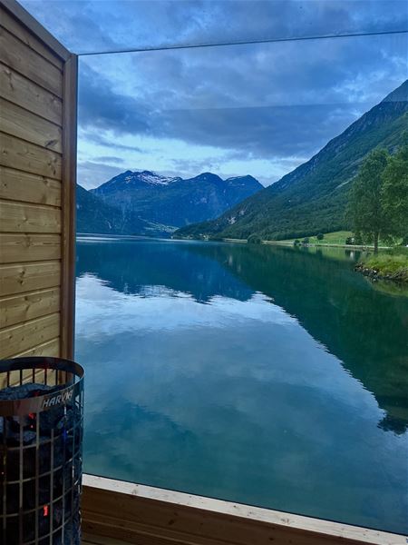 Utsikt gjennom stort glas mot roleg fjord, grøne fjell og skyer; trevegg og badstoveomn synleg til venstre -Panorama Sauna ved Strynsvatn Camping..
