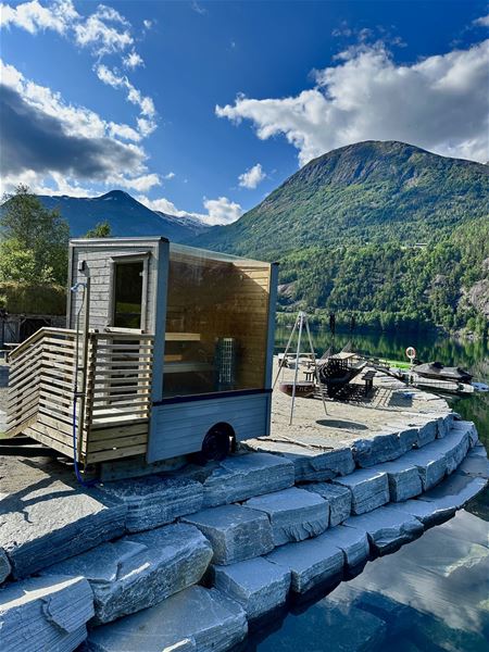 Lita badstue – Strynsvatn Camping på ei steinlagt brygge ved stille fjord, med fjell og grøne lier under blå himmel..