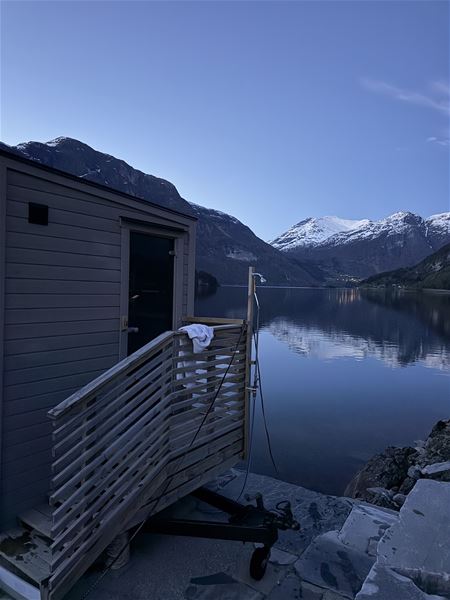 Sauna i Stryn ved ein spegelblank fjord, med snøkledde fjell og blå himmel i bakgrunnen..