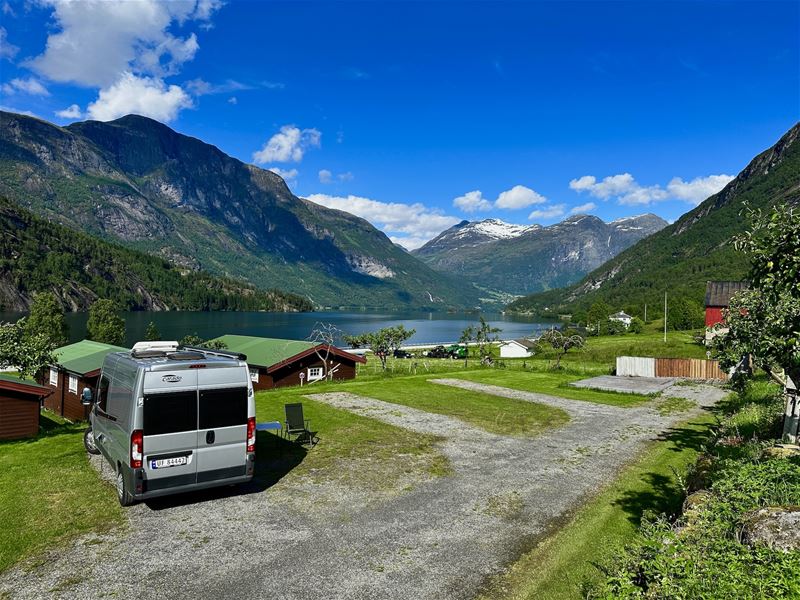 Bubil parkert på gras ved ein fjord, med hytter og bratte fjell under blå himmel med skyer..