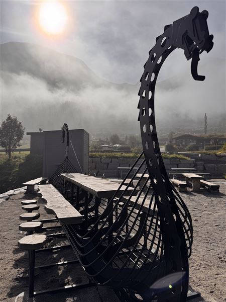 Metallskulptur av eit vikingskip ved uteservering, med sol og tåke framfor fjell i bakgrunnen i Stryn..