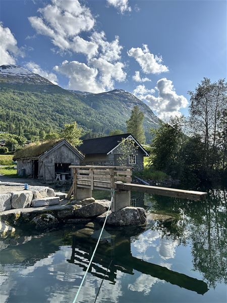 Trebygningar og ei lita bru ved eit stille vatn, med skogkledde fjell og skyer i blå himmel..