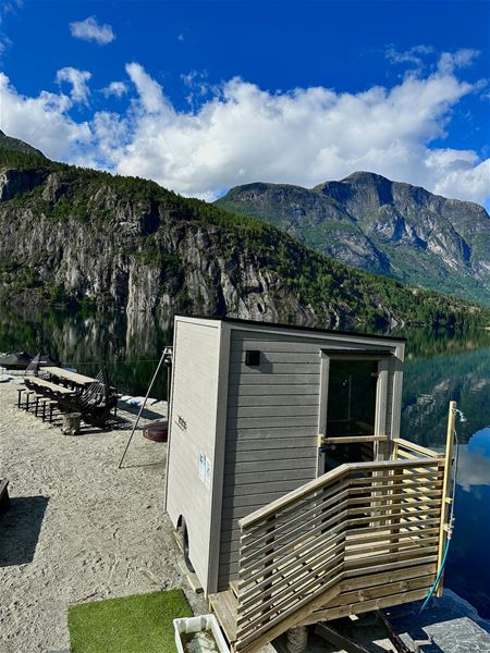 Lita badstuehytte på brygge ved stille fjord, med hengestolar, skogkledde fjell og blå himmel med skyer..