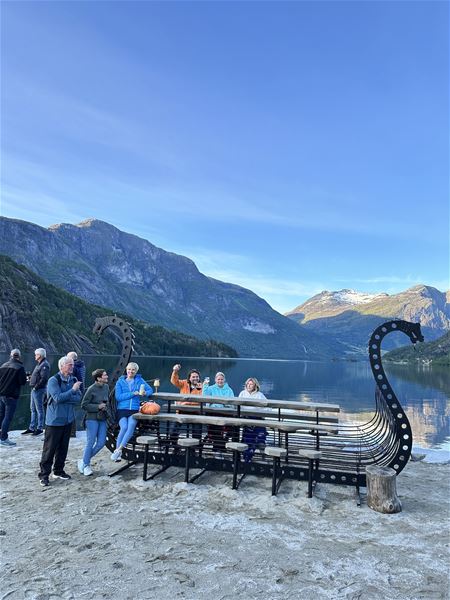 Ei gruppe personar står og sit ved ein vikingskipforma benk på sandstrand, med fjord og fjell bak..