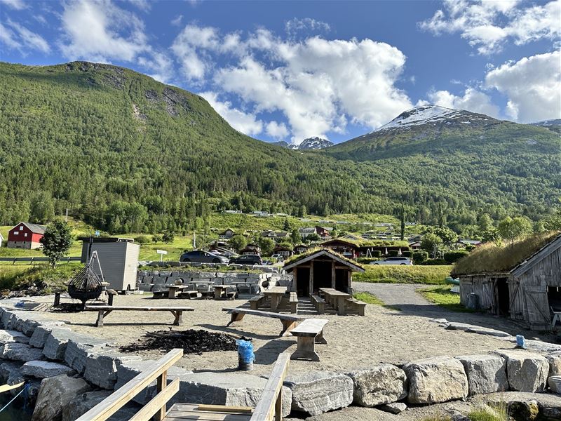 Gruslagt uteområde med benkar og bålplass framfor grøne fjell, små hus og ein snødekt topp under skyer..