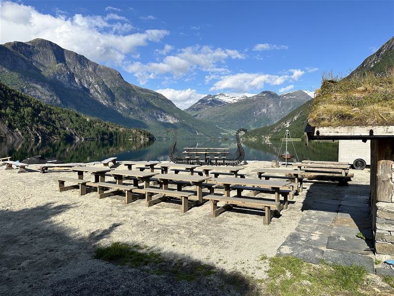 Rekkjer med trebord og benkar på ei grusflate ved roleg fjord, med bratte fjell og blå himmel..