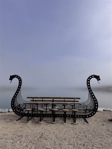 Svart drakeforma metallbenk med treplankar står på sand ved roleg vatn, med disig himmel i bakgrunnen..
