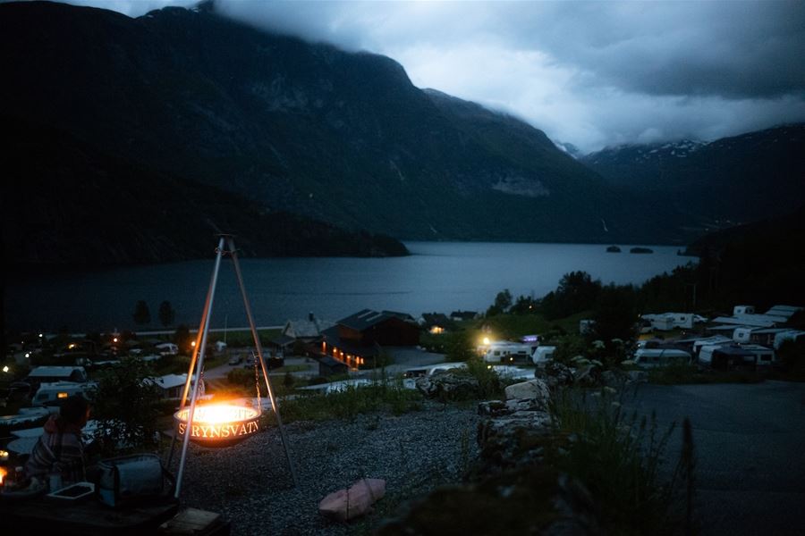 Eit glødande bål i tripod i framgrunnen, med fjord, campingplass, fjell og mørke skyer i bakgrunnen..