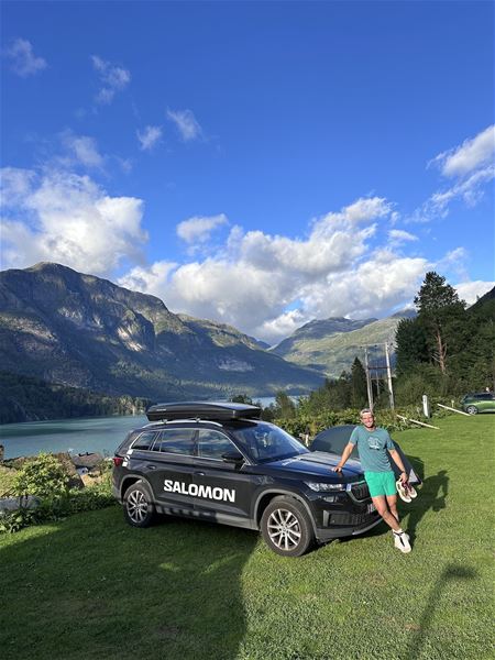 Svart stasjonsvogn på gras med takboks, ein mann ved bilen, fjord og fjell under blå himmel med skyer..
