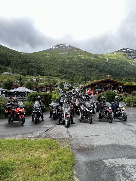 Ei stor gruppe motorsyklistar står med syklane på ein veg framfor grøne fjell og bygningar under overskya himmel..