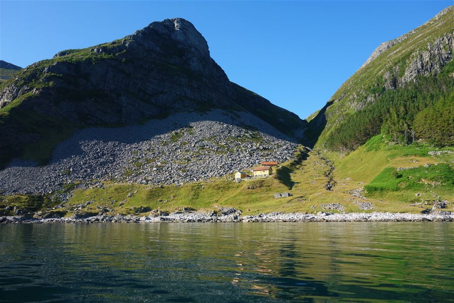 Small yellow cabin sits in a green mountain valley above calm water, with rocky slopes and a clear blue sky..