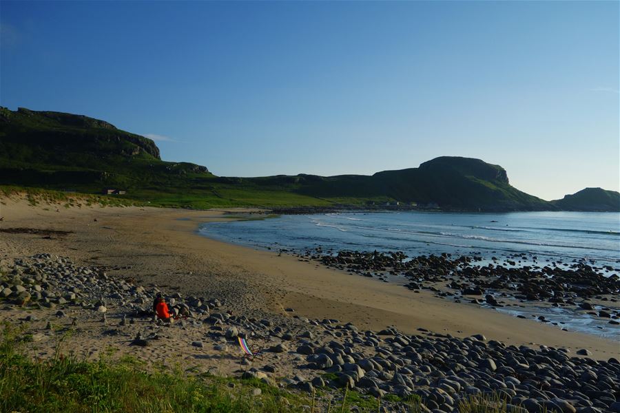 Wide sandy beach with scattered rocks, gentle waves, and green hills under a clear blue sky near sunset..