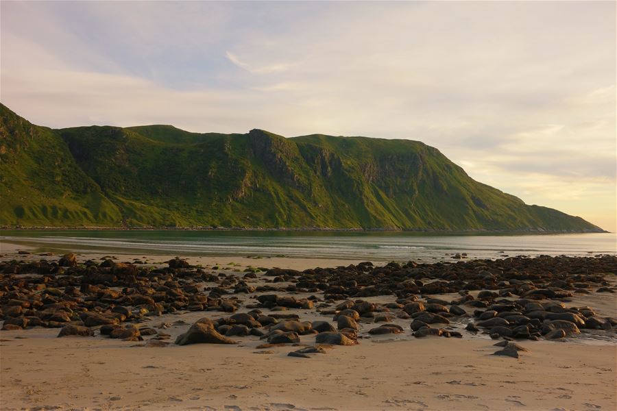 Rock-strewn sandy beach with gentle waves and a large green mountain ridge under a pale evening sky..