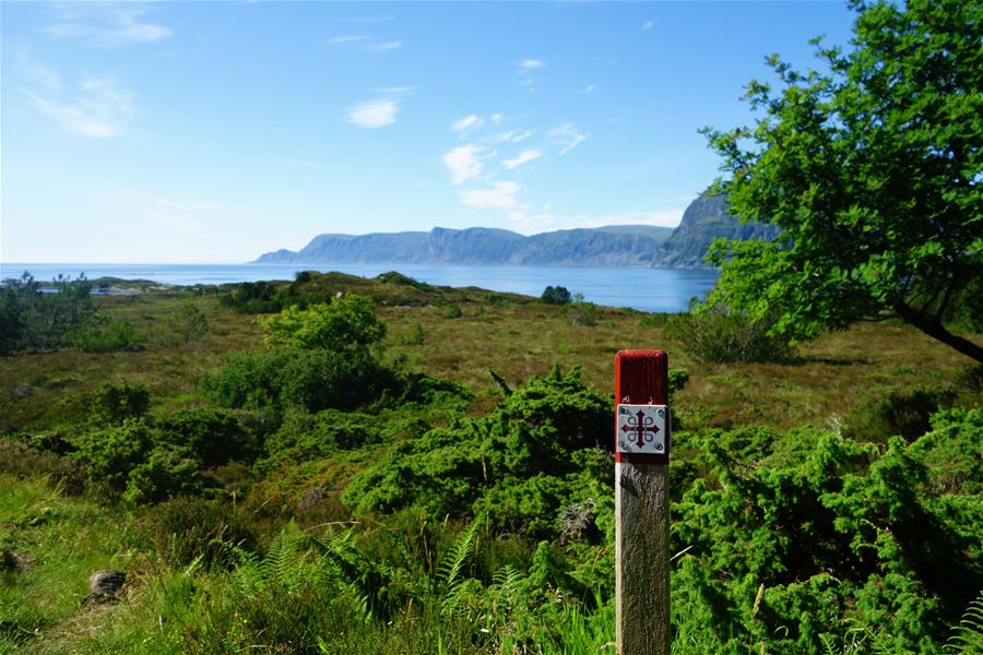 A red-topped trail marker stands in green shrubs, with a calm bay and distant mountains under a blue sky..