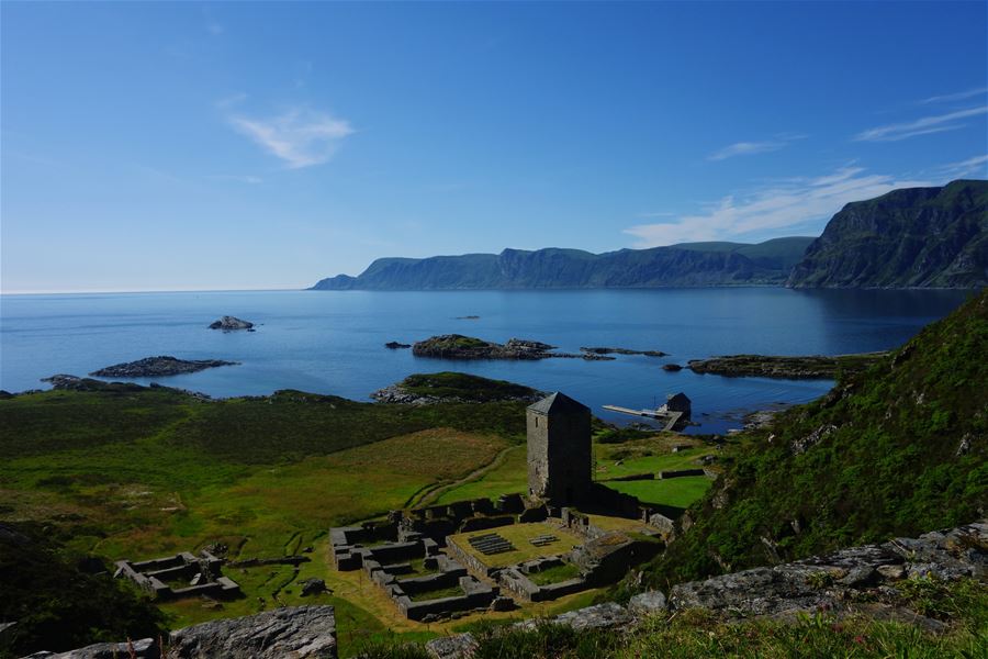 Selje Monastery – Stone tower and low ruins on a green hillside overlooking a calm blue bay with rocky islets and distant cliffs..