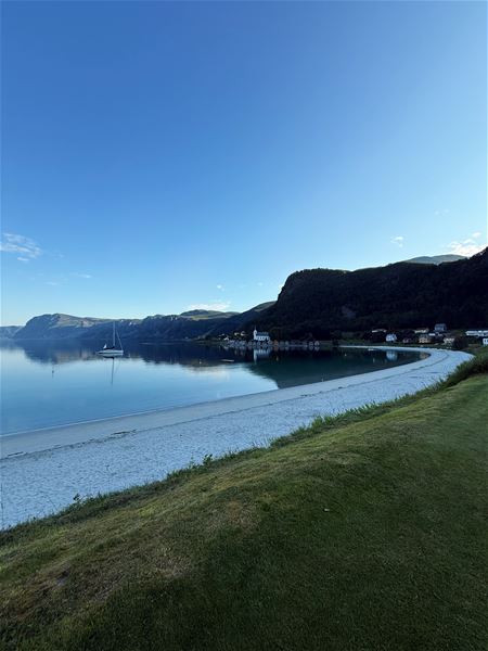 Calm bay with a sailboat near shore, curved white-sand beach, grassy foreground, and dark hills under clear sky..