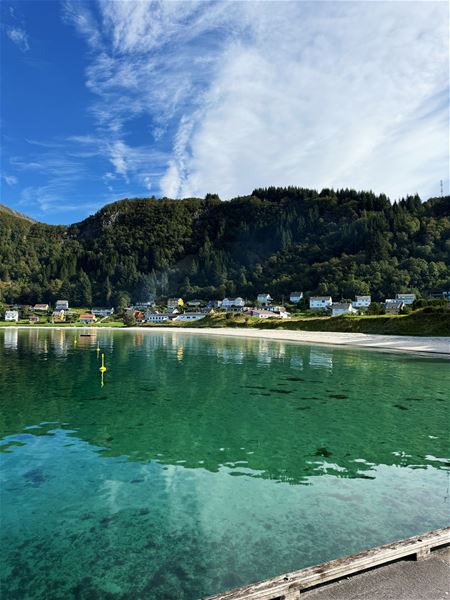Clear turquoise water in the foreground with a sandy shore, small village houses, and forested hills under a bright sky..