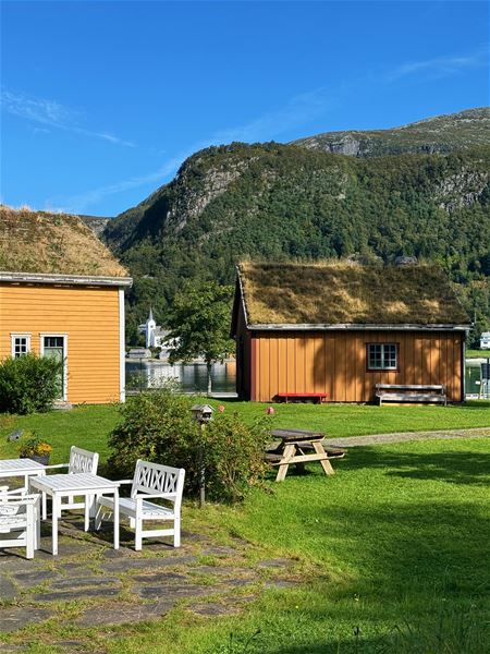 Two orange wooden cabins with grass roofs on a green lawn by a lake, with forested mountains and blue sky..