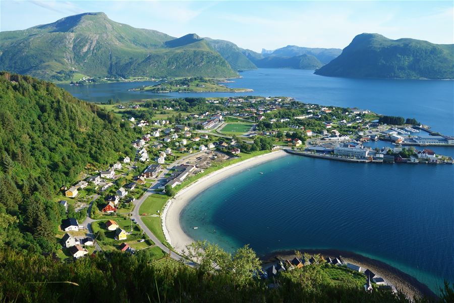 Panoramic view of a coastal village with a curved sandy beach, blue fjord, and green mountains under a clear sky..