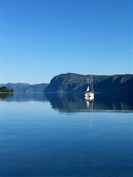 A lone sailboat floats on still blue water with a clear reflection, backed by dark green mountains under a cloudless sky..