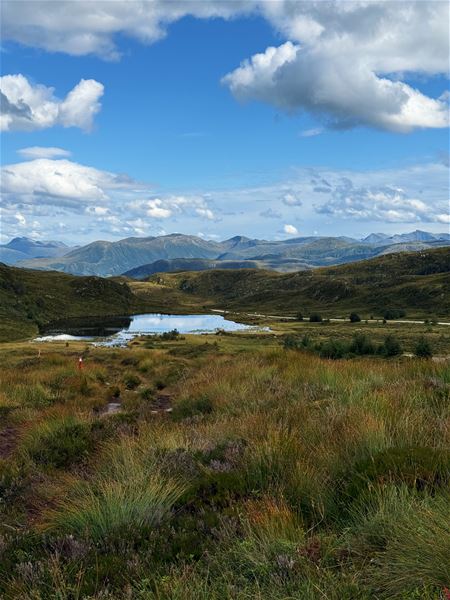 Tarvaldseggja - Topptur Ved Storhavet i Selje –Grøne fjellvidder med eit lite tjern i dalen, fjell i horisonten og blå himmel med store skyer..