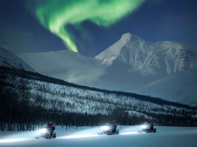 Gjester kjører snøskuter med fjell og nordlys i bakgrunnen