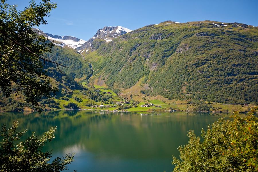Utsyn mot på Sølvene Gard Restaurant i Kandal i Gloppen. Grøne fjellsider med snøflekker ruvar over ein stille vatn, med små gardar ved strandkanten og blå himmel..