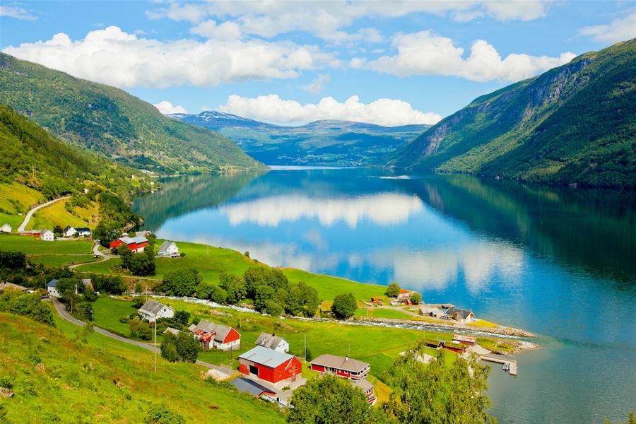 Utsyn over roleg vatn med spegling av skyer, grøne fjellsider og ein liten gard med raude hus i Kandal i Gloppen.
