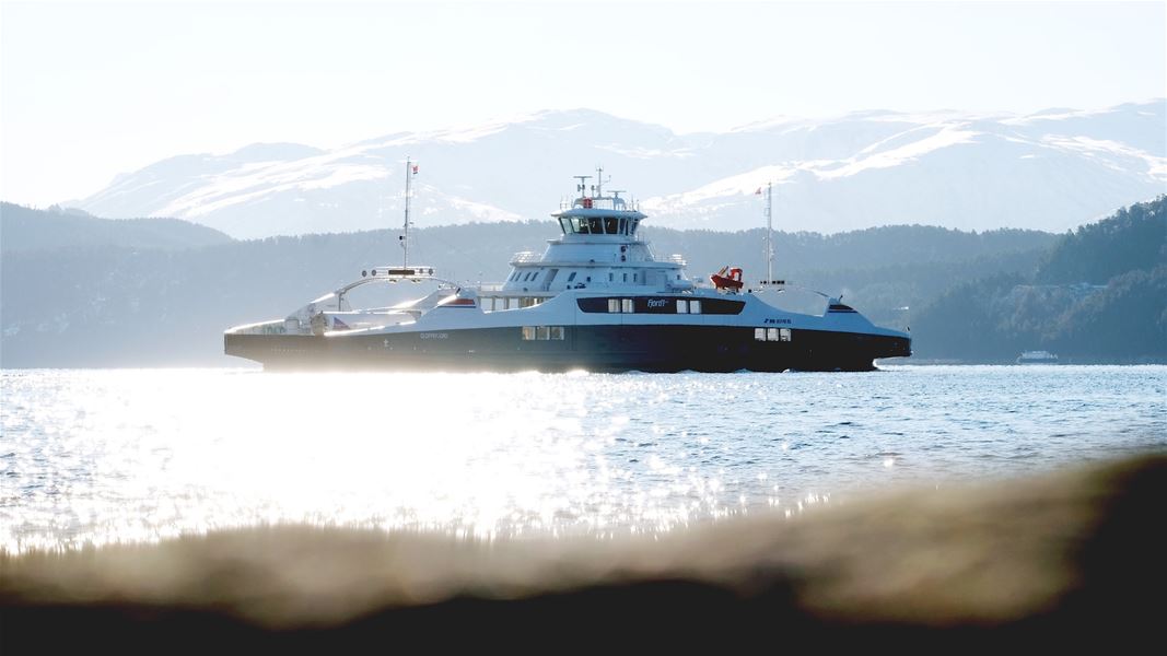 Ei blå og kvit ferje glir over ein glitrande fjord, med snødekte fjell og skogkledde åsar i bakgrunnen..