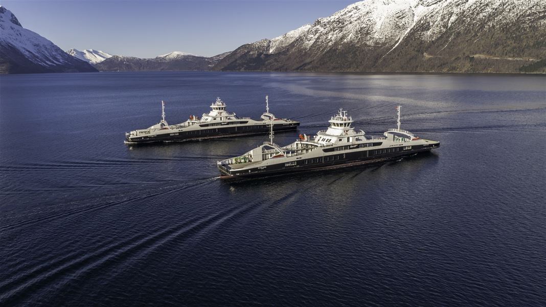 To ferjer kryssar ei roleg fjord med bølgjespor, omgitt av snødekte fjell under klar blå himmel..