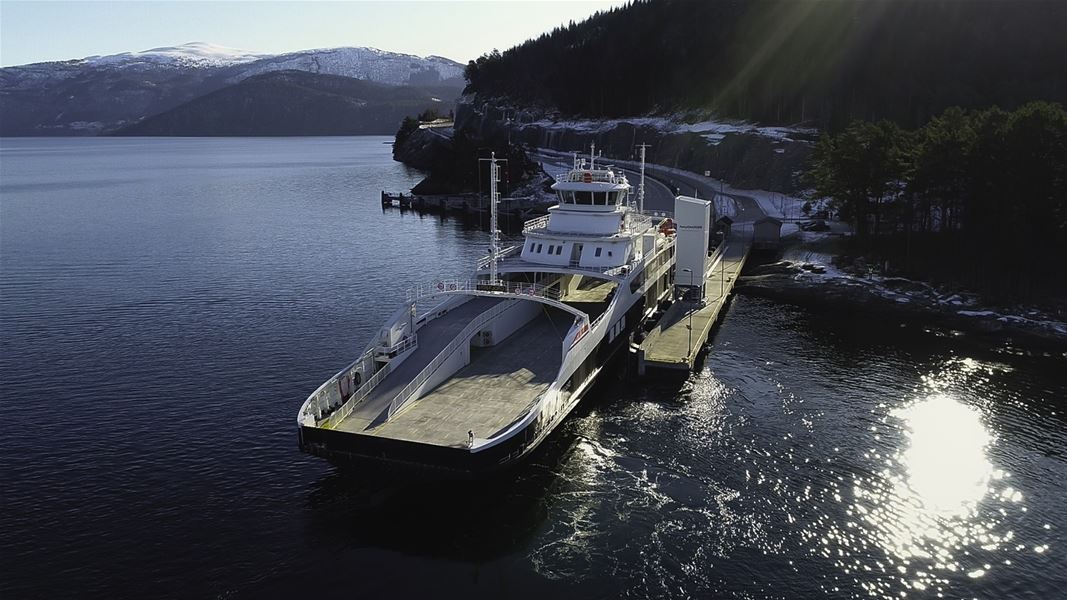 Stor bilferje ligg ved kai i roleg fjord, med skogkledd fjellside og snødekte fjell i bakgrunnen..