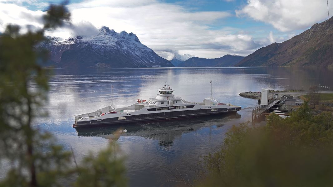 Ei bilferje ligg ved ein kai i roleg fjordvatn, med snødekte fjell og skyer i bakgrunnen..