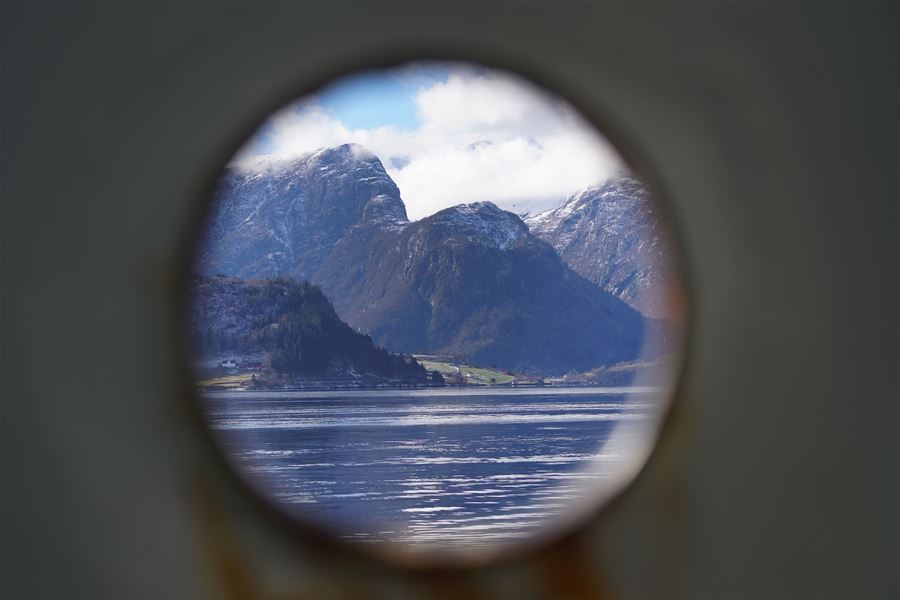 Utsikt gjennom eit rundt kikhòl mot fjord, skogkledd strand og snødekte fjell under skyer og blå himmel..