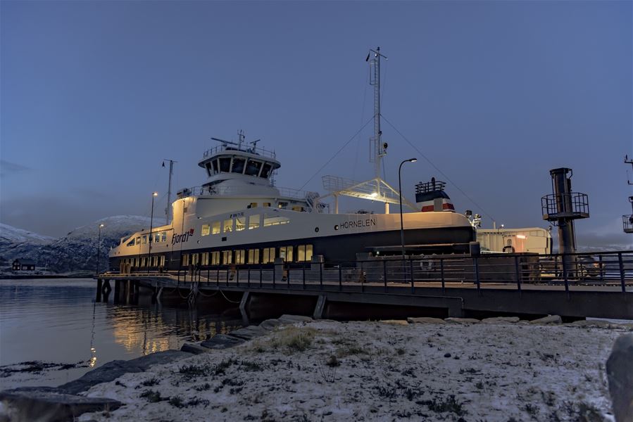 Opplyst ferje ligg ved kai i roleg fjord, med snødekt strand i forgrunnen og fjell i bakgrunnen..