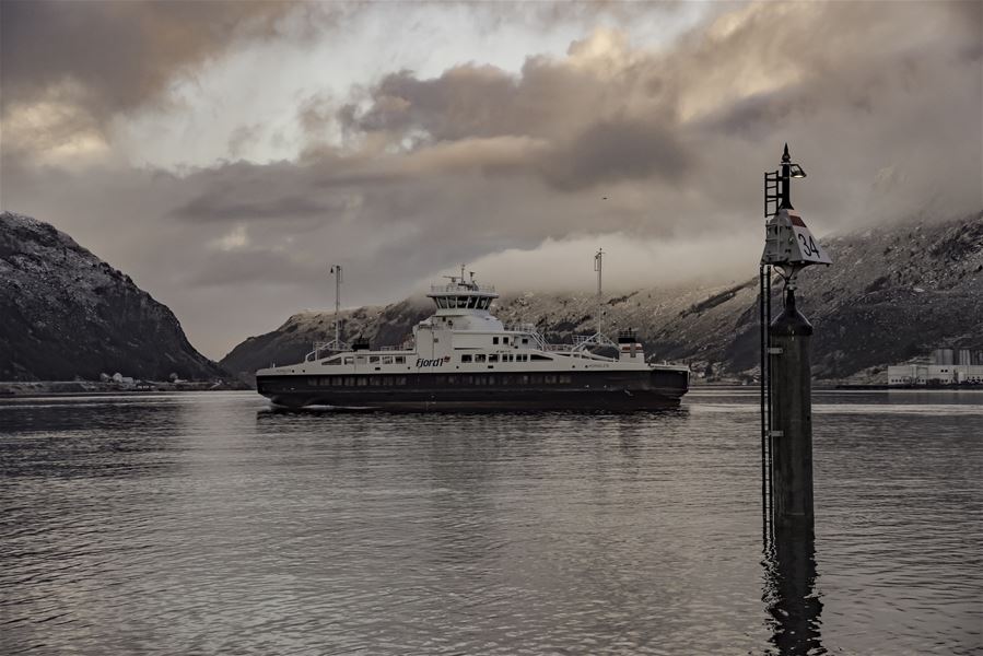 Ei ferje ligg på roleg fjordvatn mellom snødekte fjell, med ei sjømerke-stong i forgrunnen og skyer..