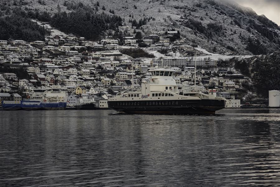 Ei stor ferje ligg på roleg fjordvatn framfor ei tettbygd, snødekt bygd i bratt fjellandskap..