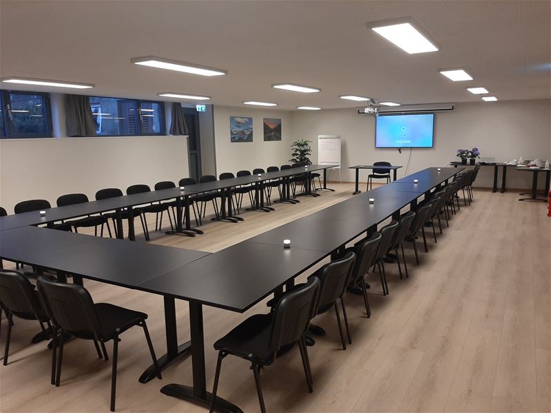 Stort møterom med U-forma svarte bord og stolar, skjerm på veggen, flippover og serveringsbord langs sida..