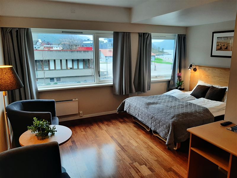 Lyst hotellrom med dobbeltseng, lenestolar, lite bord og store vindauge med utsyn mot bygningar og sjø..