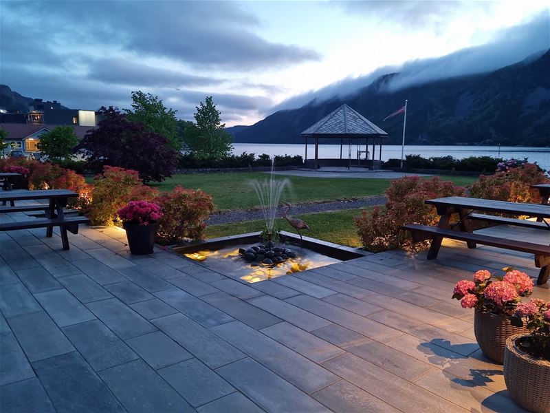 Uteplass med benkar, blomsterkrukker og liten fontene, med paviljong ved fjord og fjell i skumring..