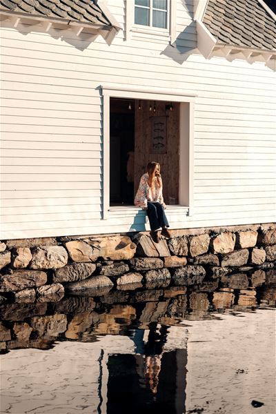 A person sits in a large open window of a white wooden building above a stone wall, reflected in calm water at the Seahouse at Hornelen Panorama in Bremanger..