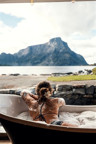 Person reclining in a bubble-filled bathtub on a patio, facing a calm lake and large mountain under clouds at the Seahouse at Hornelen Panorama in Bremanger...