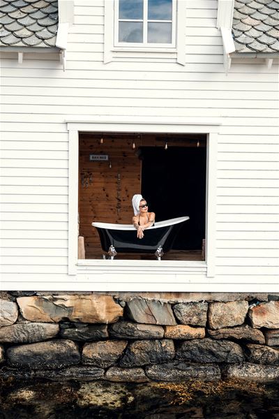 Person reclining in a black bathtub set inside a large window opening of a white beach house above a stone wall at the Seahouse at Hornelen Panorama in Bremanger..
