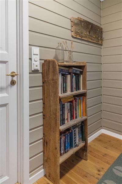 Wooden bookshelf filled with books stands by a white door on a gray paneled wall with coat hooks above at the Seahouse at Hornelen Panorama in Bremanger..